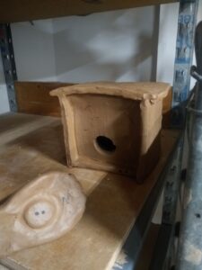 A birdhouse made out of clay. It is sitting on a piece of plywood on a shelf. A smaller mound of clay sits in front of the birdhouse.