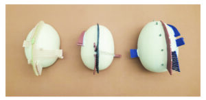 Three similar sized egg shaped forms are hanging on an orange wall. There is a central fin-like shape on all three, along with shapes jutting out on the left and right of the central fin-like shape. Thin thread that is sewn between the pieces.