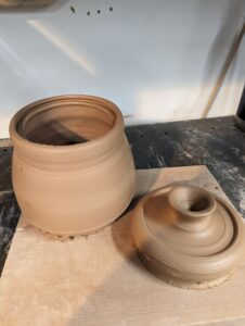 A round jar made of clay with a lid sitting next to it. Both are sitting on a piece of plywood.