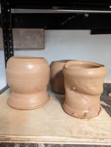 3 clay pots on a piece of plywood. The pot on the left is rounded and has a thick stem on the bottom. The pot on the right is cylindrical but twisted.