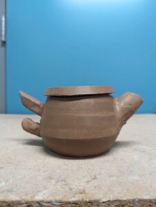 A teapot with two rods for handles made out of clay. The teapot sits on a shelf and sits in front of a blue background.