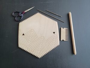 A hexagon-shaped loom, purple yarn scissors, two lengths of sewing needle, a pick-up stick, and a loom comb laid out on a black background.