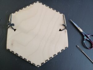 The back of the hexiloom with the warp completed, which shows how the black yarn is wrapped around the notches on the top and bottom of the loom. A sewing needle and purple yarn scissors sit beside the loom.