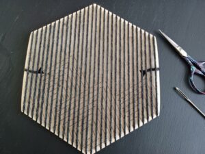 The hexiloom with the warp completed and tied off on both ends with thin black yarn. A sewing needle and purple yarn scissors sit beside the loom.