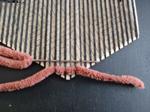 A close-up of the bottom half of the hexiloom, with warp completed and the weave started with thick, rust-coloured yarn. The thicker yarn is woven around five strands of the warp moving to the left and the needle is shown weaving around seven strands of warp moving to the right.