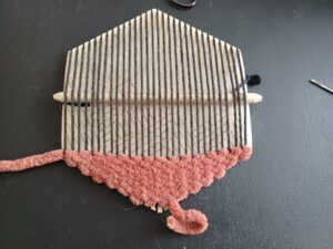 A view of the whole hexiloom with approximately a quarter of the bottom filled with the thick, rust-coloured weave. The pick-up stick is sitting underneath the warp approximately halfway up the loom.