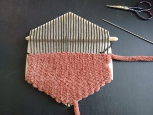 The rust-coloured weave is halfway up the hexiloom. A long sewing needle sits beside the loom, and the pick-up stick is sitting underneath the warp at the top of the loom.