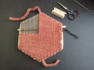The weave takes up approximately 77% of the hexiloom by filling the bottom half completely and the top half just over halfway across on the right side. The top left corner of the loom only has the thin black warp strands. The rust-coloured yarn is cut off with ends on the top and bottom, and other weaving tools sit beside the loom.