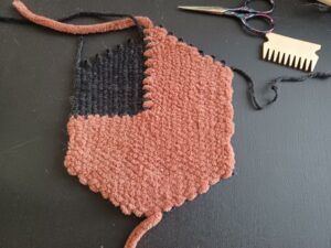 The weave now sits on a black background off of the loom. Loops made with the thin, black yarn are visible on the top and bottom of the weave where it had been wrapped around the notches of the loom. Yarn ends, two in the black yarn and two in the rust-coloured yarn, are visible coming off of the weave.