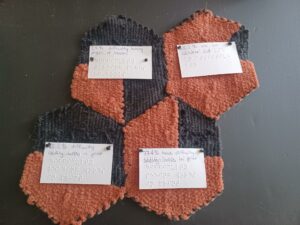 A collection of four completed weaves, seen from the back. Each has a paper label attached to the back with visual text and braille data descriptions. The first says “25% difficulty turning pages of books,” the second says “85.2% use an assistive aid,” the third says “42.4% difficulty reading words in print,” and the fourth says “77.4% have difficulty seeing words in print.”