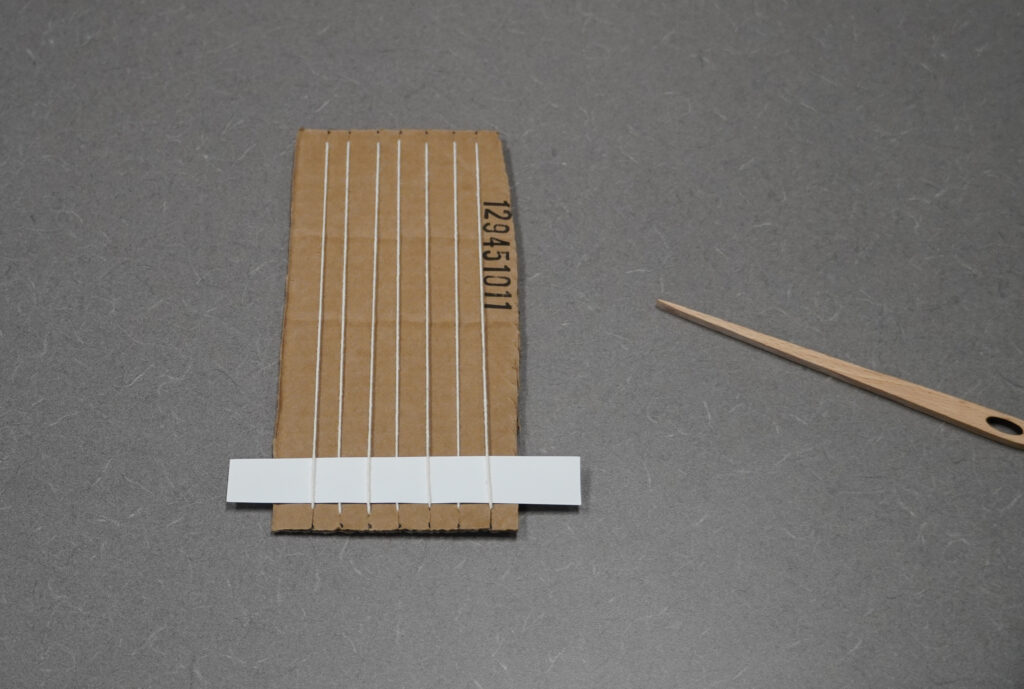 A small strip of cardstock has been woven into the bottom of the cardboard loom using plain weave. 