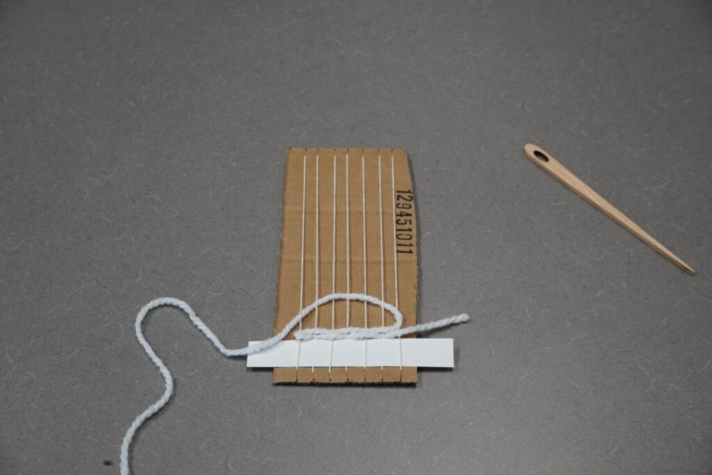 A few lines of plain weave (over and under) are on the loom. The weft is pulled up in an arch across the warp to be lightly pushed down against the other threads.