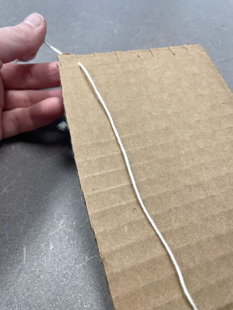 A cardboard loom. A strip of cardboard has been cut into a makeshift loom. The top and bottom of the cardboard have a series of shallow cuts at regular half-inch intervals. A string is being nestled into the first notch at the top of the cardboard. 