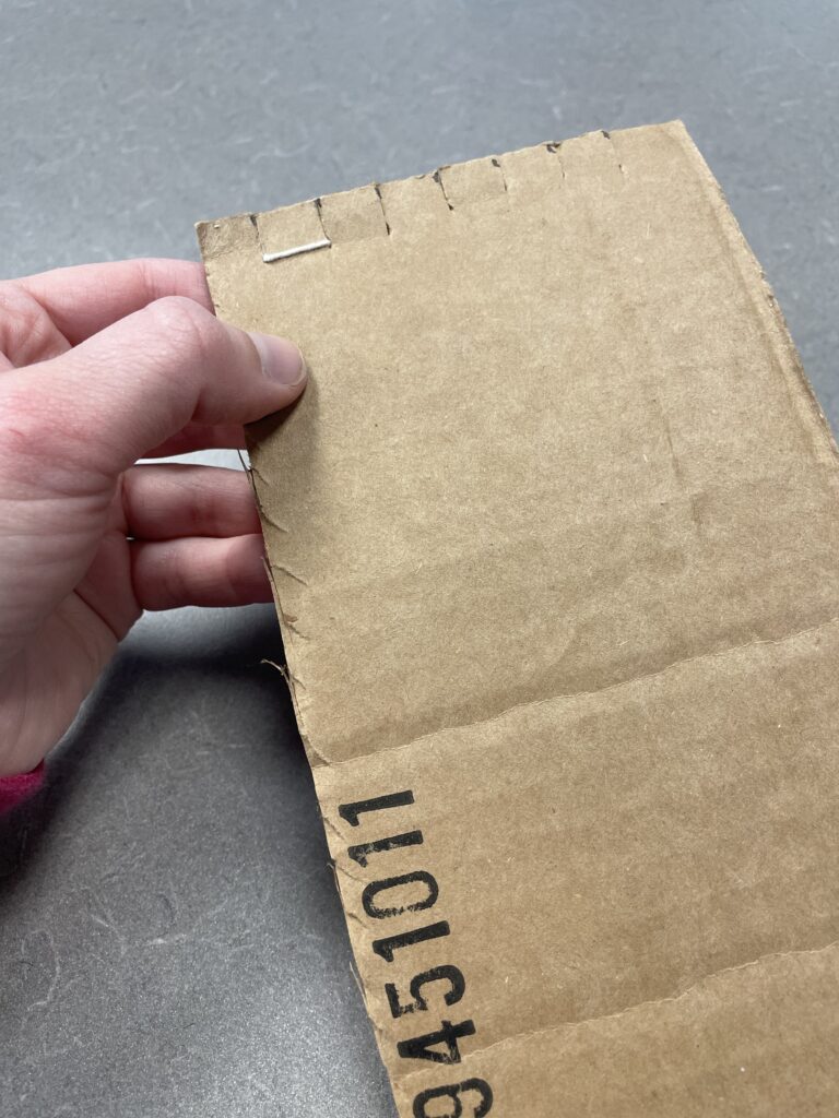 The back side of the cardboard loom. One string has been put on and the second is started. From the back, the only string visible is a horizontal stitch between two notches.