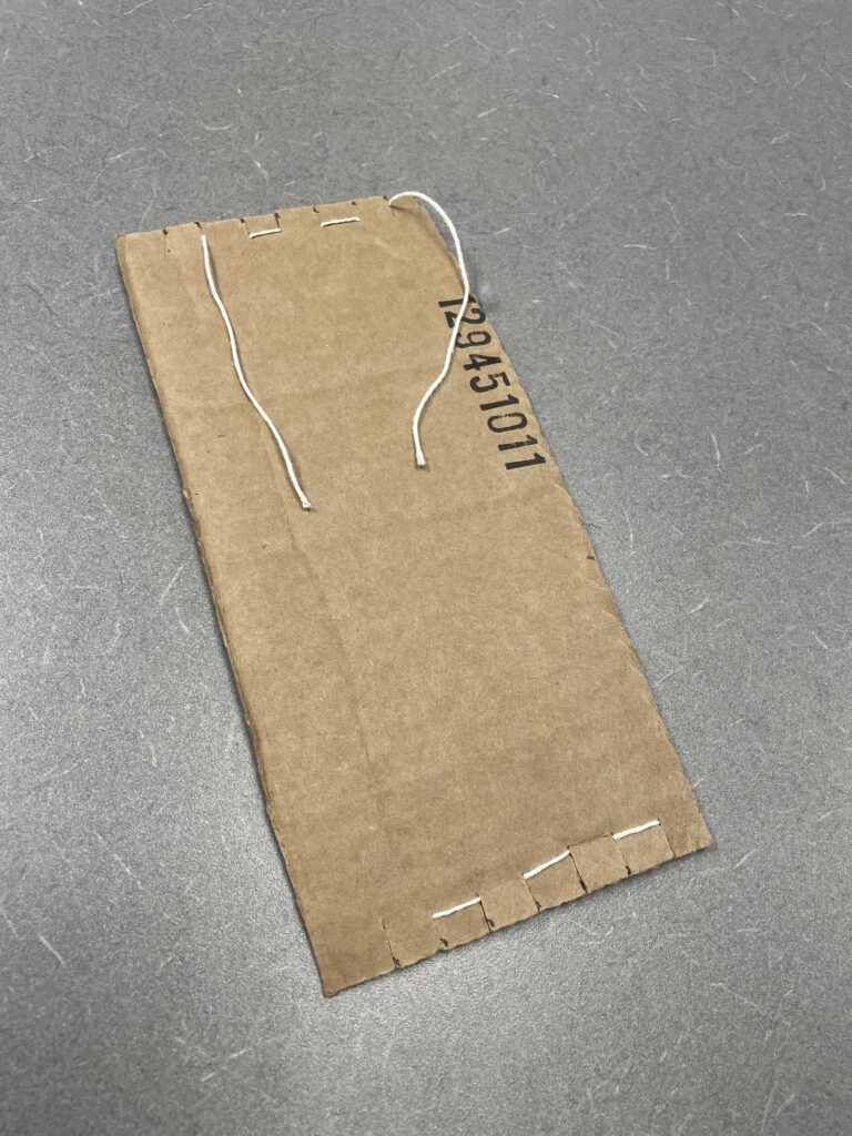 A completed cardboard loom from the back. The only string that is visible are the horizontal stitches between notches which alternate between top and bottom. The string starts and ends on the same side and long tails have been left.