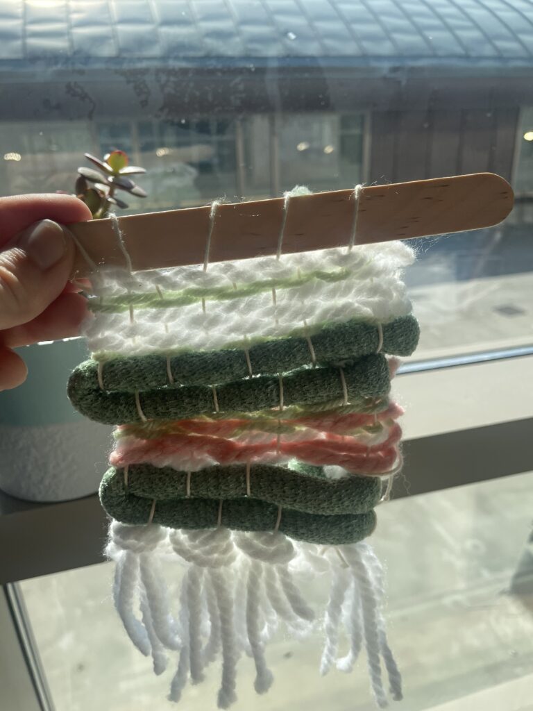 A miniature tapestry of the CDC website, strung up on a popsicle stick, held up to a window with light streaming in beside it.